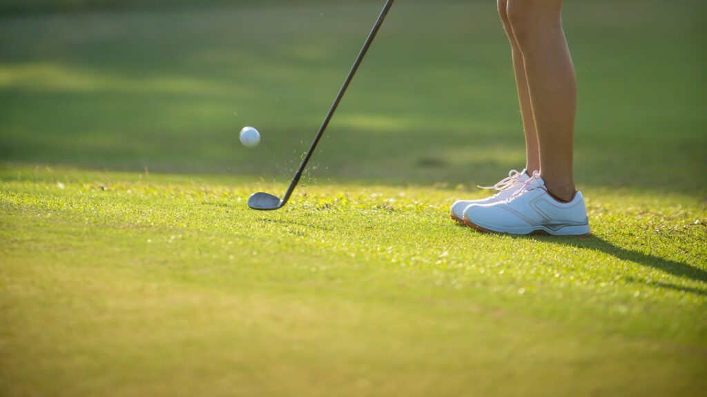Legs and white golf shoes of a person about to hit a golf ball with a golf club on a green golf course.